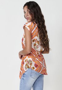 Top de patrón floral multicolor con mangas cortas, con acentos en naranja, rojo y verde sobre un fondo blanco, combinado con vaqueros de color azul claro.