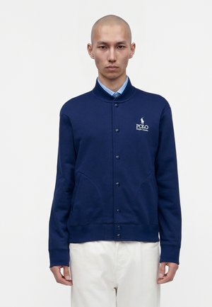 Mann trägt eine marineblaue, geknöpfte Polo Ralph Lauren Jacke über einem hellblauen Hemd und weißen Hosen, stehend vor einem schlichten weißen Hintergrund.