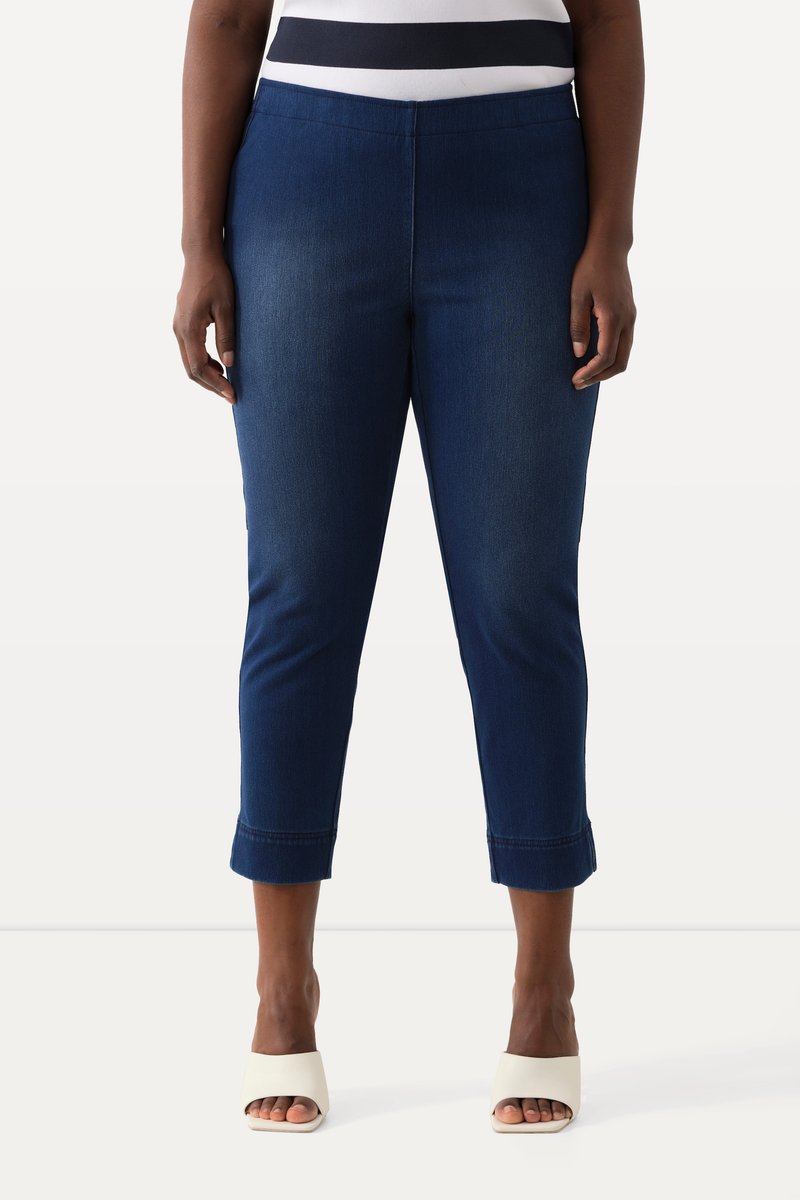 Kvinde iført mørkeblå cropped denim bukser og hvide åbne hæle, står mod en ensfarvet lys baggrund.