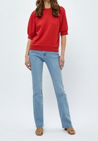 Žena oblečená v červeném topu s nadýchanými rukávy, světlých modrých rovně střižených džínech a béžových sandálech na blokovém podpatku s otevřenou špičkou, stojící na jednobarevném pozadí.
