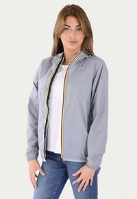 Letvægts grå jakke med hætte, gul og sort lynlåsdetalje, elastiske manchetter og logo på venstre bryst. Kombineret med en hvid top.