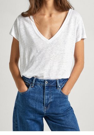 Frau trägt weißes V-Ausschnitt-Kurzarmshirt, das in hochgeschnittene blaue Jeans gesteckt ist, mit Händen in den Taschen vor einem schlichten Hintergrund.