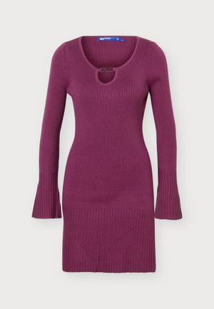 Femme debout portant une robe en maille côtelée violette ajustée à manches longues et des bottes en cuir noires jusqu'au genou sur un fond clair.
