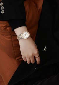 Reloj redondo de plata y oro rosa con un dial texturizado, marcadores de gemas, unido a una pulsera de metal. Mano descansando sobre una tela marrón.