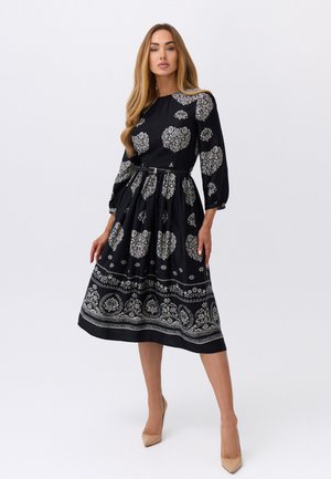 Kobieta ubrana w czarną sukienkę midi z białym kwiatowym wzorem, długimi rękawami, paskiem podkreślającym talię oraz beżowe szpilki na białym tle.