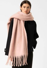 Mujer con una gran bufanda suave de color rosa claro con flecos, drapeada sobre una chaqueta negra, de pie frente a un fondo claro y liso.