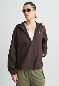 Καφέ ελαφρύ μπουφάν με κουκούλα, κλείσιμο με φερμουάρ και λευκό λογότυπο στο αριστερό στήθος. Φοριέται με λευκό αμάνικο τοπ και π ράσινο παντελόνι.