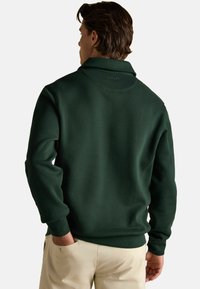 Mężczyzna w ciemnozielonym, długim swetrze z kołnierzykiem, stojący z jedną ręką w kieszeni beżowych spodni, zwrócony do nas plecami.