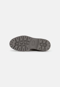 Semelle de chaussure marron avec des crampons profonds et de petits motifs géométriques, présentée sur un fond gris clair uni.