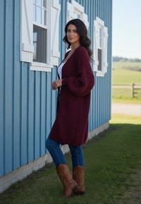 Burgundowy oversize kardigan z teksturowanym splotem, z kieszenią, w połączeniu z niebieskimi dżinsami i brązowymi kowbojskimi butami na tle niebieskiej ściany.