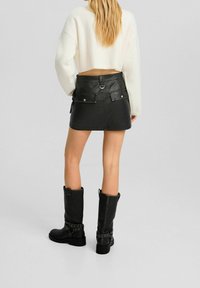 Svart läder minikjol med framfickor, silveraccenter och bältesöglor. Matchad med knähöga svarta stövlar med spänndetaljer.