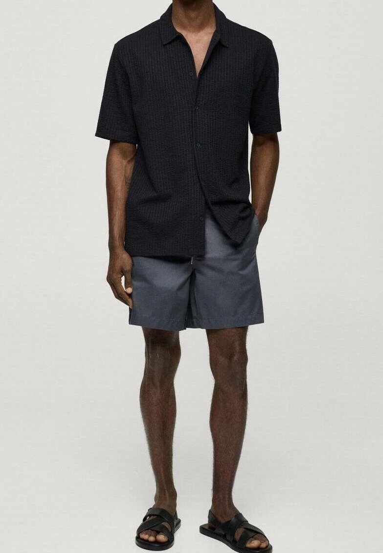 Homme portant une chemise noire à manches courtes boutonnée, un short gris et des sandales noires, debout avec une main dans la poche.