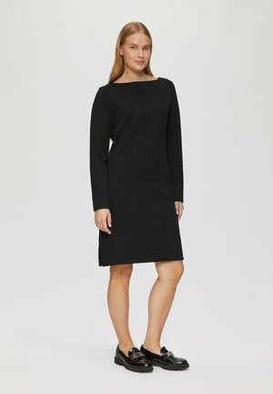 Robe noire à manches longues avec un décolleté bateau, motif chevrons texturé, longueur genou, fentes latérales, associée à des mocassins noirs brillants.