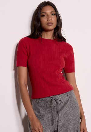 Femme portant un haut tricoté rouge texturé à manches courtes et un pantalon gris à cordon, debout devant un fond clair uni.