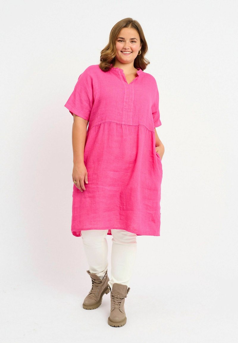 Abito tunica di lino rosa con maniche corte, scollatura a V e vita arricciata. Abbinato a pantaloni bianchi e stivaletti beige alla caviglia.