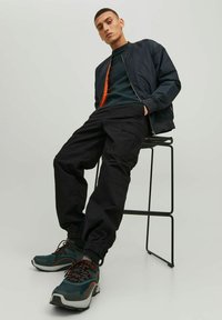 Giubbotto bomber nero sopra un maglione verde scuro, abbinato a pantaloni cargo neri e sneakers teal con accenti arancioni. Seduto su uno sgabello di metallo.