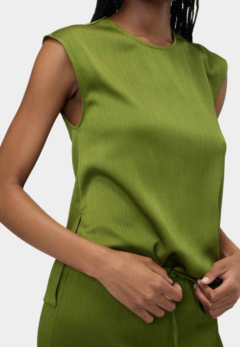 Femme aux cheveux foncés tressés portant un haut sans manches texturé vert olive, ajustant le tissu près de la taille.