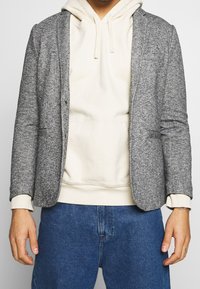 Grauer strukturierter Blazer über einem cremefarbenen Hoodie mit aufgesetzten Taschen und legerer Passform, kombiniert mit blauem Denim.