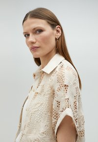 Lace shirt in cream with floral pattern, short sleeves, and a button-up design. Notable cream collar and metallic gold earrings.