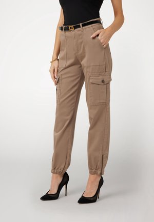 Mujer con pantalones cargo beige con puños elásticos, tacones altos negros y cinturón negro con hebilla dorada, con la mano en el bolsillo, sobre fondo liso.