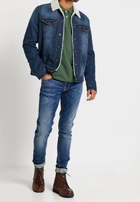 Jeansjacka med lammullskrage, grön polo skjorta, blekta blå jeans och bruna snörstövlar, med uppvikta ärmar på jeansen.