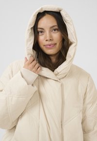 Beige pufferjakke med stor hætte, quiltet tekstur og ribbede manchetter. Har en central spændeåbning og en afslappet pasform.