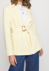 Yellow linen blazer with a belted waist, flap pockets, notched lapels, and long sleeves, paired with a white t-shirt and blue jeans.