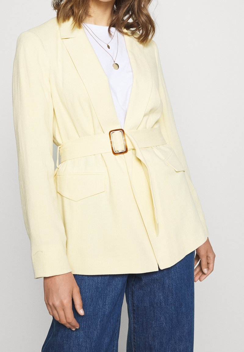 Yellow linen blazer with a belted waist, flap pockets, notched lapels, and long sleeves, paired with a white t-shirt and blue jeans.