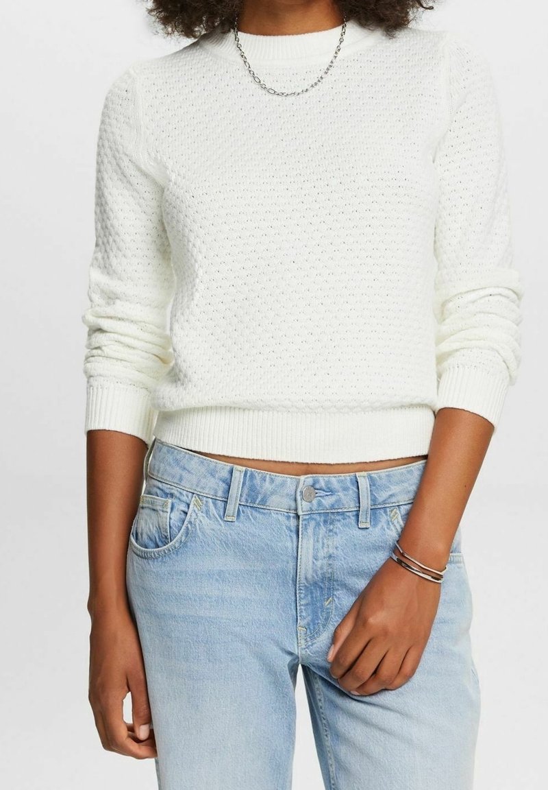 Femme portant un pull en maille blanche texturée, un jean taille haute bleu clair, un collier chaîne argenté et des bracelets au poignet droit.