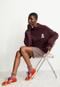 Bordeaux trui met opstaande kraag, geborduurd lintdetail; bruine rok; rode sneakers met blauwe veters, grijze sokken. Zittend op een chroom stoel.