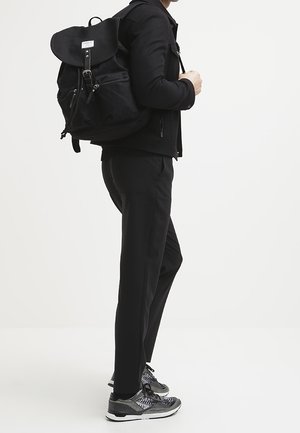 Černý plátěný batoh s vrchním klopovým víkem, přední kapsou na zip a nastavovacími popruhy, který nese osoba v černých kalhotách a se vzorovanými teniskami.