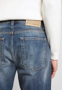 Denimjeans i medelblå tvätt med en mjuk textur, försedd med en tan läderetikett med perforeringar och kontrasterande sömmar på fickorna.