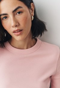 Pink ribbed crewneck top with a rounded neckline, paired with gold textured earrings. Smooth skin with subtle freckles in a close-up portrait.