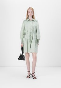 Hellgrünes Kleid mit Kragen, Knopfleiste und Kordelzug in der Taille. Mit langen puffartigen Ärmeln mit Raffung. Kombiniert mit einer schwarzen Tasche.