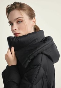 Kobieta z jasnymi włosami w czarnej kurtce puchowej, trzymająca kołnierz blisko twarzy, lekko spoglądająca na bok.