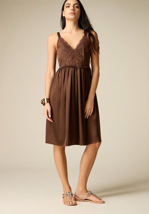 Vrouw draagt een mouwloze bruine jurk met kanten afwerking en geplooide rok, gouden manchetarmband en platte sandalen met kralen, staand tegen een beige achtergrond.