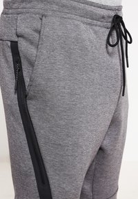 Gråa joggers med dragsko i midjan, sidofickor med dragkedja och ett texturerat tyg som kombinerar mjuka och släta element.