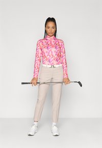 Golf apģērbs, kurā ir rozā garā piedurkne augšdaļa ar ziedu rakstu, bēšas piegrieztas bikses un baltās sporta apavi, turot golfu nūju.