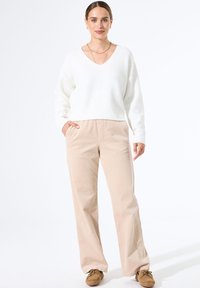 Vrouw staat met hand in zak, gekleed in een witte V-hals trui, beige wijde broek, bruine schoenen en gouden sieraden voor een witte achtergrond.