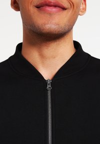 Giacca nera con zip, caratterizzata da un colletto a coste, una texture liscia e un dettaglio in metallo sulla zip frontale, che mette in risalto un design minimalista.