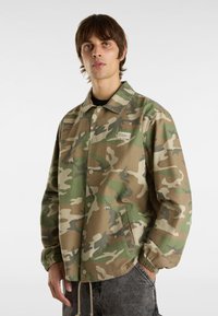 Tarnmusterjacke in Grüntönen und Beige, aus strapazierfähigem Material, mit Druckknopfverschluss, Seitentaschen und elastischen Bündchen.