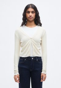 Κρεμ ριμπ πλεκτή ζακέτα με μακριά μανίκια, κλείσιμο με κουμπιά και κοντό σχέδιο. Συνδυασμένη με σκούρα μπλε τζιν ψηλόμεση.