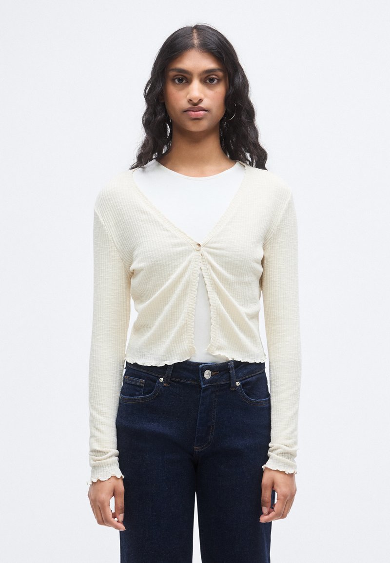 Κρεμ ριμπ πλεκτή ζακέτα με μακριά μανίκια, κλείσιμο με κουμπιά και κοντό σχέδιο. Συνδυασμένη με σκούρα μπλε τζιν ψηλόμεση.