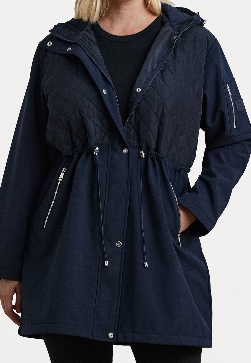Navy blue jacket featuring a quilted upper section, drawstring waist, dual zippered pockets, and a detachable hood. Soft fabric texture.