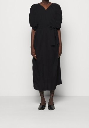 Kobieta w czarnej, plisowanej sukience midi z bufkami na rękawach, z paskiem w talii i czarnych, płaskich sandałach na paski na jednolitym białym tle.