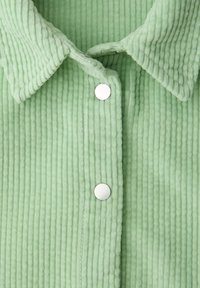 Veste en velours côtelé vert clair avec un col pointu et deux boutons-pression ronds argentés visibles sur le devant.