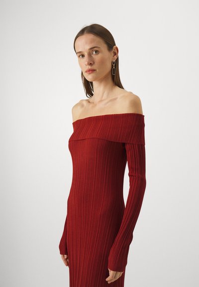 Femeie cu păr drept, brunet, îmbrăcată cu o rochie roșie, cu mâneci lungi, tip off-shoulder, din material texturat, purtând cercei geometrici din aur, privindu-și înainte.