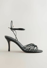 Sandale noire à talons hauts avec une fine bride à la cheville et plusieurs fines lanières sur l’orteil ouvert sur un fond neutre.