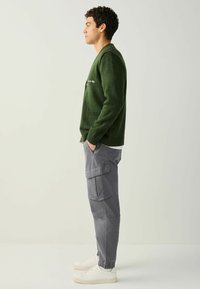 Homme portant un cardigan vert, un pantalon cargo gris et des baskets blanches, debout de profil, les mains dans les poches, sur un fond clair.