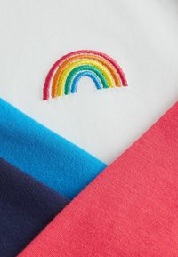 Multikolorni materijal na bijeloj pozadini sadrži vez u obliku duge. Vidljivi su teksturirani dijelovi u crvenoj, plavoj i mornarsko plavoj boji.